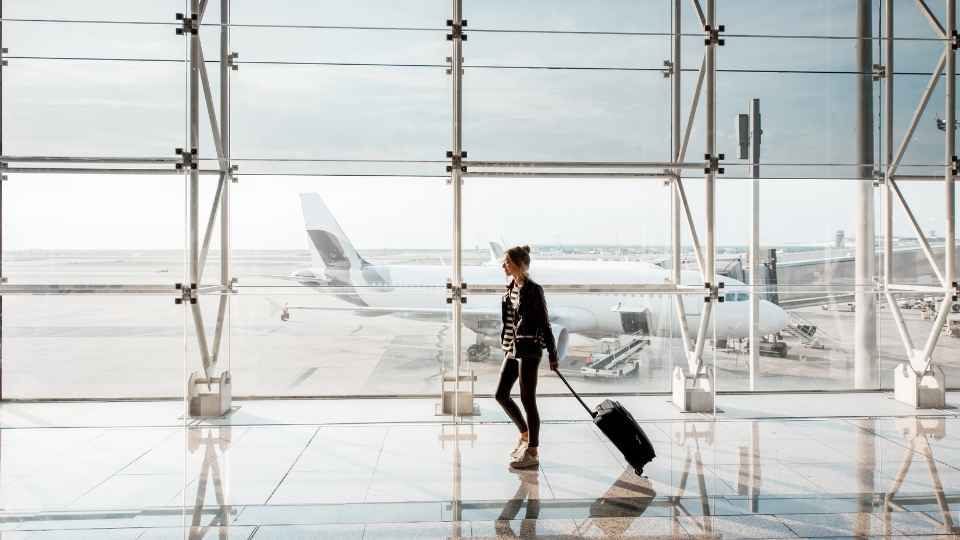 A passenger walking through an airport terminal with a suitcase, with a plane preparing for departure outside the window.
