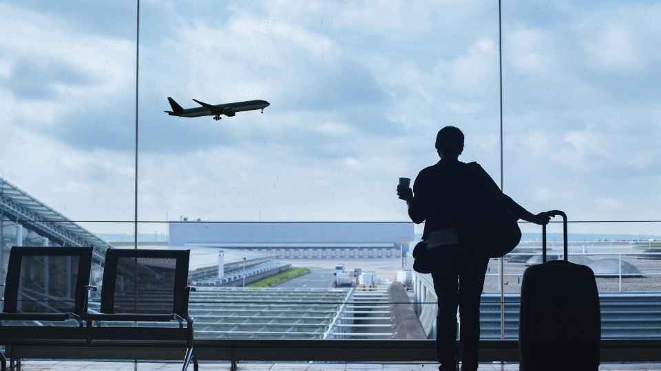 A traveler standing in an airport terminal with luggage, watching a plane depart through the large window.