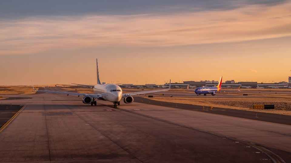 Two airplanes taxiing on the runway during sunset, with the airport terminal visible in the distance.