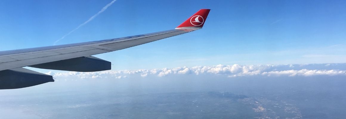 Airplane wing high above land and coastline, with a clear blue sky and a line of clouds on the horizon.